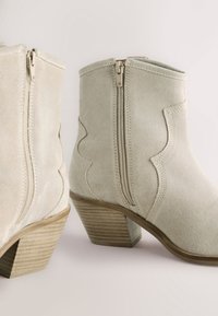 Beige suède enkellaarsjes met een zigzagpatroon, ritssluitingen en een gestapelde houten hak. Gladde textuur en een ronde neusvorm.