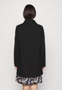 Cappotto in lana nera con design sartoriale, maniche lunghe e ampio colletto, indossato sopra una gonna a fantasia con dettagli floreali.