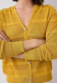 Gelbes Strickcardigan mit einem strukturierten Design in unterschiedlichen Breiten, V-Ausschnitt und einer einzelnen Knopfverschluss vorne. Gerippte Bündchen.