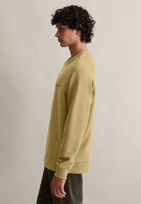 Lichtgele crewneck sweatshirt met ribbels aan de manchetten en onderkant, met een kleine accent op de borst. Model draagt een donkere corduroybroek.