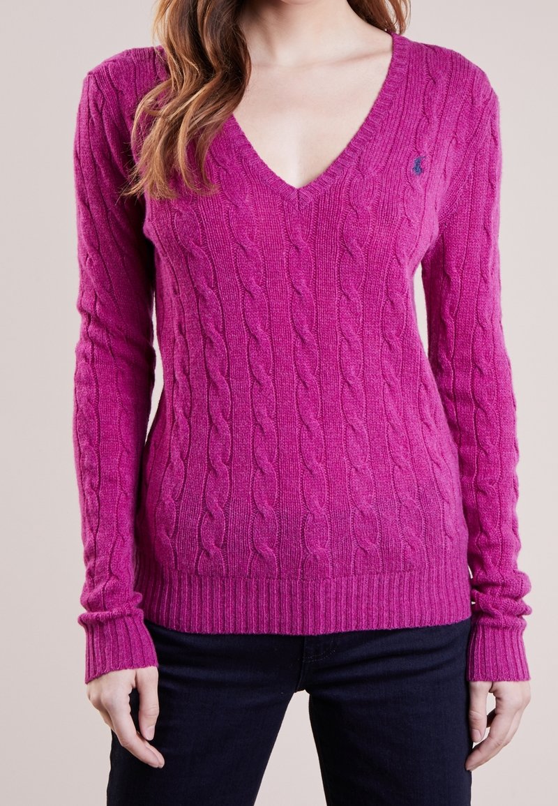 Femme portant un pull à manches longues, en maille torsadée, col en V magenta et un pantalon sombre, vue du cou aux hanches sur un fond neutre.