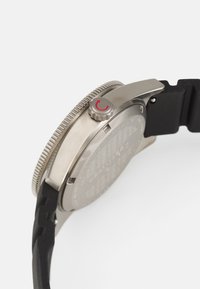 Montre en acier inoxydable avec une lunette grise, un bracelet en caoutchouc noir et une couronne proéminente avec un accent rouge. Le dos montre des détails gravés.