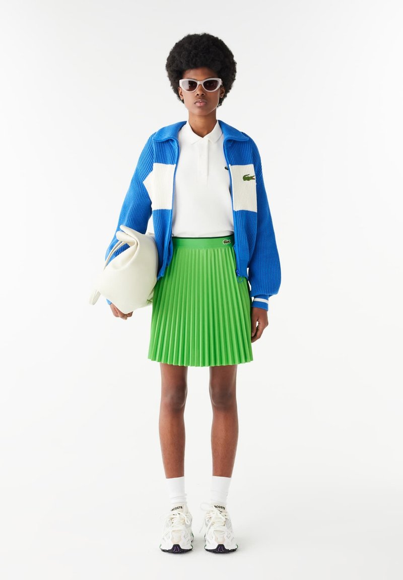 Niebieska kurtka z zamkiem, z prążkowanym wzorem i białymi akcentami, biała koszulka polo, zielona spódnica z fałdami oraz białe tenisówki z czarnymi akcentami. Jasne tło.