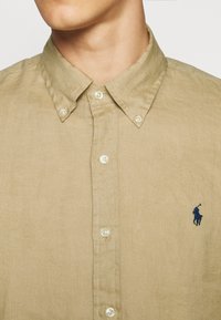 Beige košile z lněného materiálu s knoflíky, button-down límcem a tmavě modrým vyšitým logem polohokejisty na levé hrudi.
