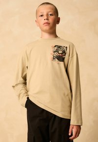 Beige long-sleeve t-shirt featuring a graphic of a gorilla, round neckline, relaxed fit, paired with black pants. Smooth cotton texture.