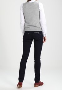 Ljusgrå ärmlös tröja över vit coll shirt, kombinerad med svarta jeans och vinröda skor. Slät textur och figursydd design.