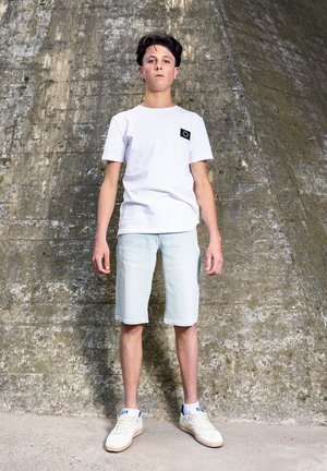 Teenager-Junge steht vor einer strukturierten Betonwand, trägt ein weißes T-Shirt, helle Jeansshorts, weiße Turnschuhe und weiße Knöchelsocken.