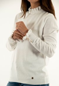 Maglione bianco con un colletto alto e arricciato, texture a pois e dettagli con bottoni perle sui polsini. Presenta un orlo a costine e maniche lunghe.