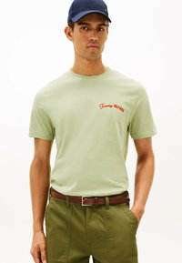 Lysegrønt bomulds t-shirt med rødt "Tommy Hilfiger" logo, korte ærmer og rund hals. Sæt sammen med olivengrønne bukser og et brunt bælte.