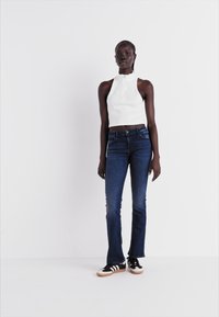 Haut sans manches blanc avec col montant, jeans évasés bleu foncé, et chaussures de sport noires avec des accents blancs et beiges. Design simple et ajusté.
