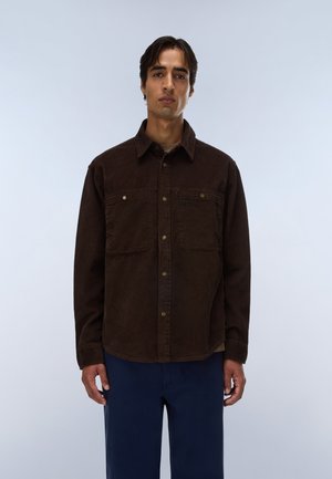 Giovane uomo con capelli corti e scuri indossa una camicia di velluto a coste marrone scuro con bottoni e pantaloni blu scuro, in piedi davanti a uno sfondo chiaro e semplice.