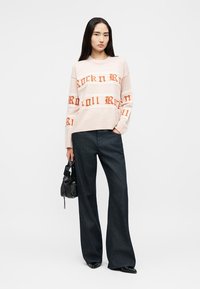 Γυναίκα που φοράει ανοιχτό μπεζ πουλόβερ με πορτοκαλί κείμενο «Rock n Roll», φαρδιά σκούρα μπλε παντελόνια, μαύρα παπούτσια και κρατά μια μικρή μαύρη τσάντα.