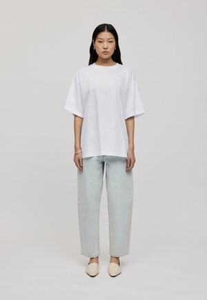 White oversized cotton t-shirt with short sleeves, paired with light blue loose-fitting jeans and beige slip-on shoes. Plain background.