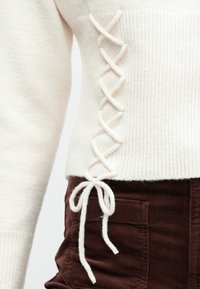 Close-up de um suéter de malha creme com detalhe de laço lateral amarrado em forma de laço sobre o bolso de calças de veludo cotelê castanho-escuro.