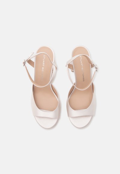 Paire de sandales blanches à bout ouvert avec bride à la cheville, à talons bas, posées à plat sur un fond clair, montrant des semelles intérieures beiges.