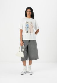 White graphic t-shirt featuring two stylized figures in blue and pink. Paired with gray baggy shorts, white sneakers, and a small white handbag.