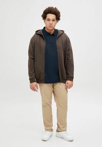 Brūns zip-up kapucis virs tumši zila polo krekla, apgūts ar bēšiem biksēm un baltām kedām. Ikdienišķa apģērba kompozīcija ar mīkstām tekstūrām.