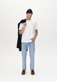 Bijela pamukasta majica s logom, svijetlo plave traperice, smeđe cipele i tamni beanie, s crnom jaknom prebačenom preko ramena.