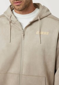 Bežinė zip-up gobtuvė su minkšta, zomšai panašia tekstūra, reguliuojamais raišteliais ir ryškiu "GUESS" logotipu šviesiai tan spalvos ant krūtinės.