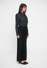 Denim overhemd met knoopsluiting aan de voorkant, borstzakken en contrasterende stiksels, gecombineerd met hoge zwarte wijde broek en zwarte flats.