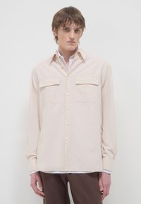Camicia beige a maniche lunghe con chiusure a bottoni, due tasche sul petto e una texture morbida; indossata sopra una camicia bianca con colletto.