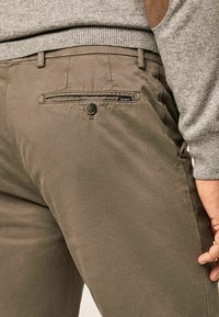 Detailní záběr na osobu v hnědých kalhotách Hackett s knoflíkovou zadní kapsou a svetrem z lehkého šedého úpletu.