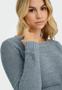 Femme blonde portant un pull en laine bleu clair, tenant l'encolure avec sa main droite, expression faciale neutre, fond blanc.