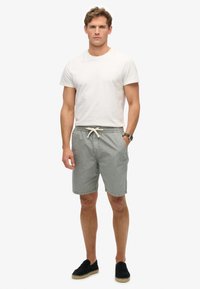 Shorts gris clair pour homme avec une taille élastique et un cordon de serrage. Associés à un t-shirt blanc ajusté. Chaussures noires à enfiler. Design minimaliste.