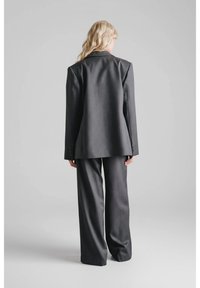 Kobieta z długimi blond włosami ubrana w oversize'owy ciemnoszary garnitur, widziana od tyłu na tle jednolitego jasnoszarego tła.