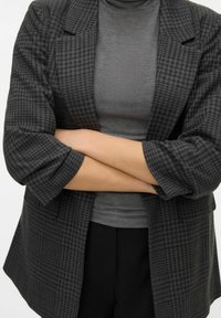 Gråternet blazer med løs pasform og oprevne ærmer, båret over en grå turtleneck. Stoffet ser tekstureret ud med subtile mønstre.