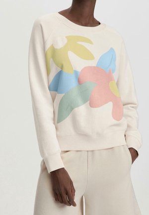 Persoon draagt een crèmekleurige trui met oversized pastel bloemenmotief in geel, blauw, roze en groen, gecombineerd met bijpassende crèmekleurige broek.
