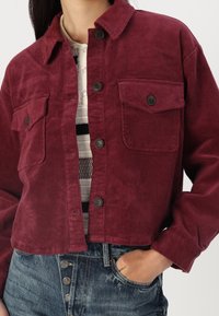 Corduroyjakke i dyb burgunder med sorte knapper, med to brystlommer og et cropped design. Bæres over en stribet skjorte.