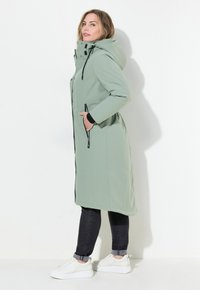 Cappotto impermeabile lungo di colore verde chiaro con cappuccio, tasche con zip nere e taglio dritto. Il modello indossa jeans neri e sneakers bianche.
