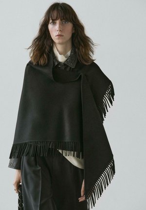 Frau mit schulterlangem braunem Haar, die einen schwarzen Fransenschal über einer schwarzen Lederjacke und schwarzen Hosen vor einem einfarbigen Hintergrund trägt.