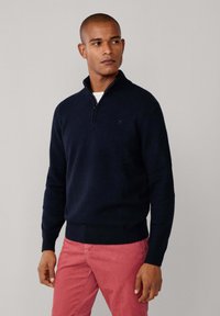 Marineblau gestrickter Pullover mit einem Reißverschlusskragen. Rippbündchen und -saum, mit einem dezenten Logo auf der Brust. Kombiniert mit rosa Hosen.
