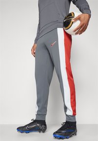 Hombre con pantalones deportivos grises, blancos y rojos sostiene un balón de fútbol blanco y negro, vistiendo botines negros con suelas azules.