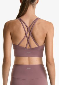 Mauve sportbeha met gekruiste rugbanden, naadloos ontwerp en OYSHO-branding in zilver boven de elastische band. Textuur van de stof afwerking.