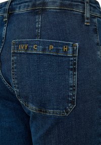 Vaqueros de mezclilla en azul oscuro con un acabado texturizado. Presentan un bolsillo trasero con el texto en relieve "IVY C P H" en color beige.