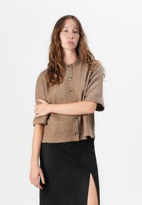 Brun kortermet cardigan laget av mykt materiale med rund hals, med gullknapper og en avslappet passform, kombinert med et svart skjørt.