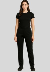 A woman stands confidently, wearing a black t-shirt and black pants with decorative side stripes, against a neutral gray background.