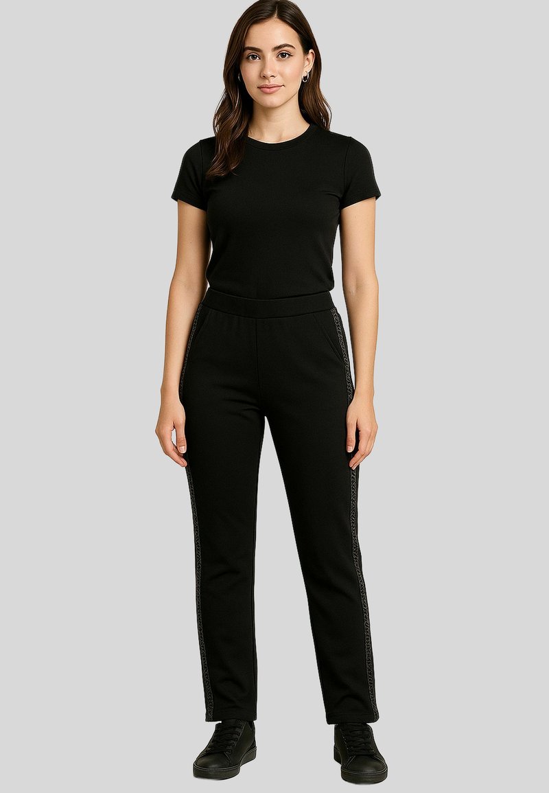 A woman stands confidently, wearing a black t-shirt and black pants with decorative side stripes, against a neutral gray background.