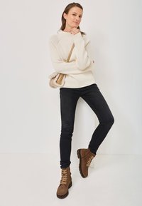 Maglione beige a coste, jeans skinny neri e stivali in suede marroni con lacci. Una piccola borsa beige con bordi morbidi completa l'outfit.