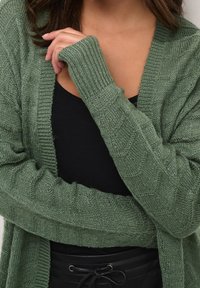 Zielony dzianinowy kardigan z prążkowanymi mankietami i teksturowanym wzorem, noszony na czarnej bluzce. Materiał wydaje się miękki i przytulny.