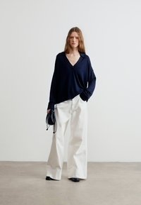 Γυναίκα που φοράει χαλαρό μαύρο πλεκτό με V λαιμόκοψη, φαρδιά άσπρη παντελόνια, μαύρα παπούτσια, κρατώντας μια μικρή μαύρη τσάντα, στέκεται σε απλό πάτωμα μπροστά σε λευκό τοίχο.