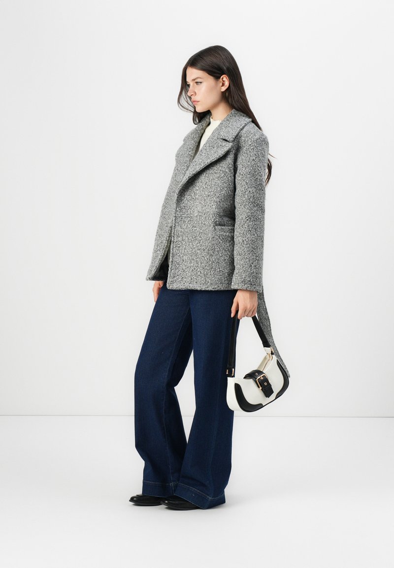 Manteau en laine texturé gris avec un col cranté, poches latérales, associé à un pantalon large bleu marine et un petit sac à main noir et blanc.