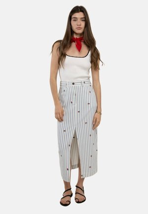 Mujer con cabello largo y castaño que lleva una camiseta blanca sin mangas, pañuelo rojo en el cuello, falda midi a rayas con bordado floral y sandalias negras.
