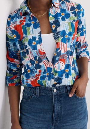 Personne portant une chemise bleue à imprimé floral boutonnée sur un haut blanc rentré dans un jean bleu taille haute avec une main dans la poche.