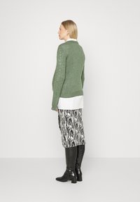 Suéter verde de malha com um padrão texturizado, sobre uma camisa branca, combinado com uma saia com padrão preto e branco e botas de cano alto pretas e brilhantes.