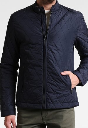 Homme portant une veste matelassée bleu marine zippée avec col cheminée et poches latérales zippées, une main dans la poche, associée à un pantalon vert olive.