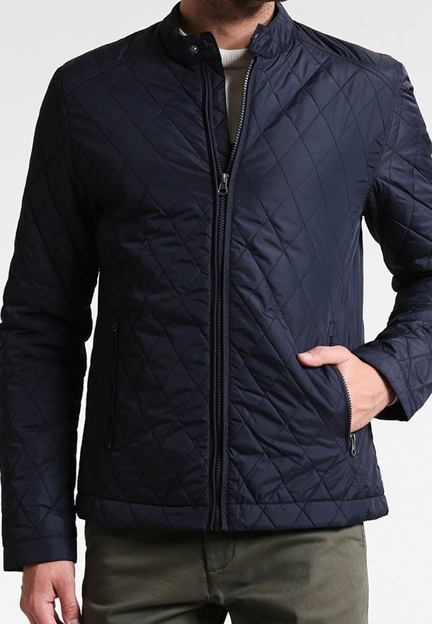 Homme portant une veste matelassée bleu marine zippée avec col cheminée et poches latérales zippées, une main dans la poche, associée à un pantalon vert olive.